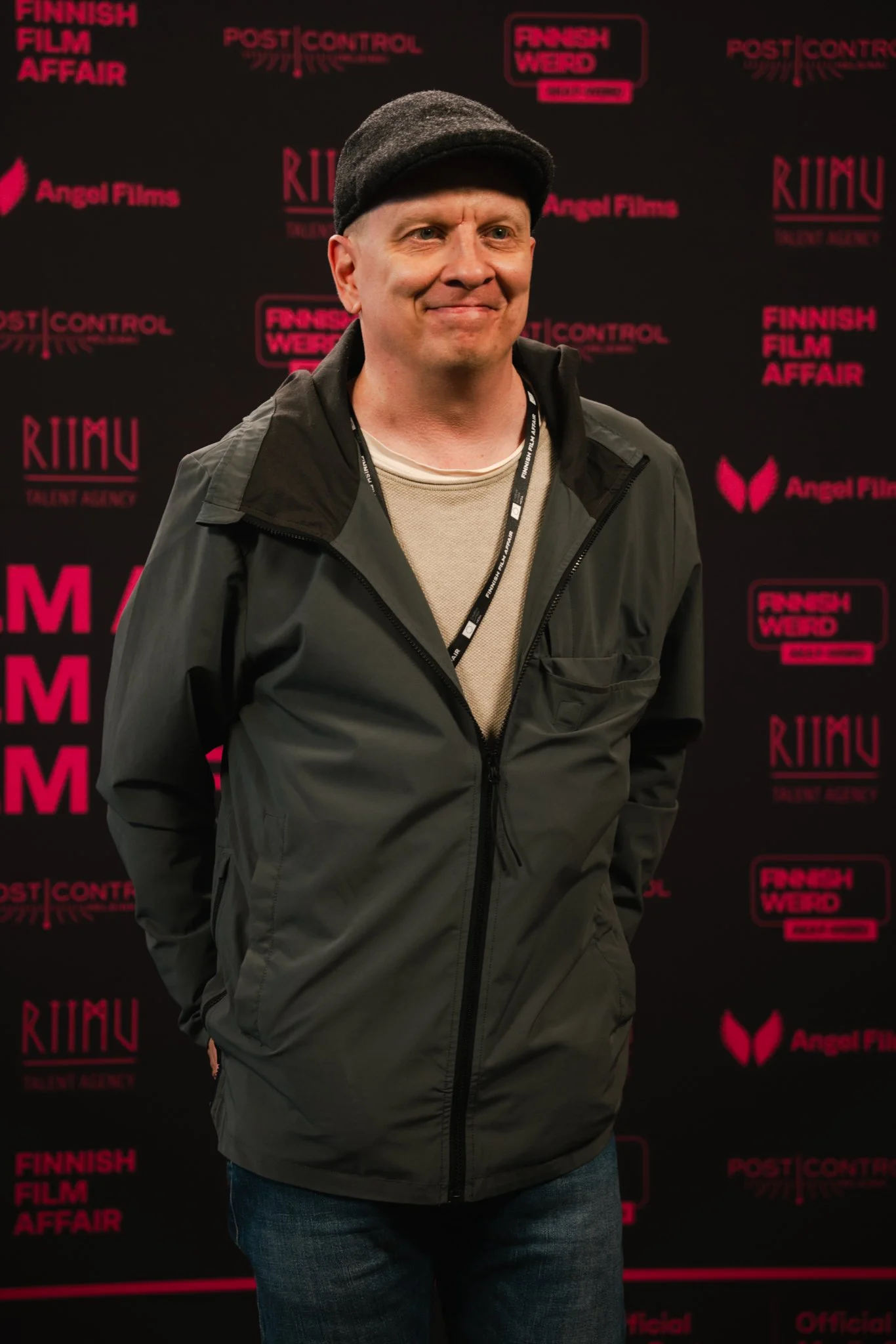 A man in a black jacket and beanie standing in front of a black backdrop with pink and white logos and text related to the Finnish Film Affair and other film organizations.