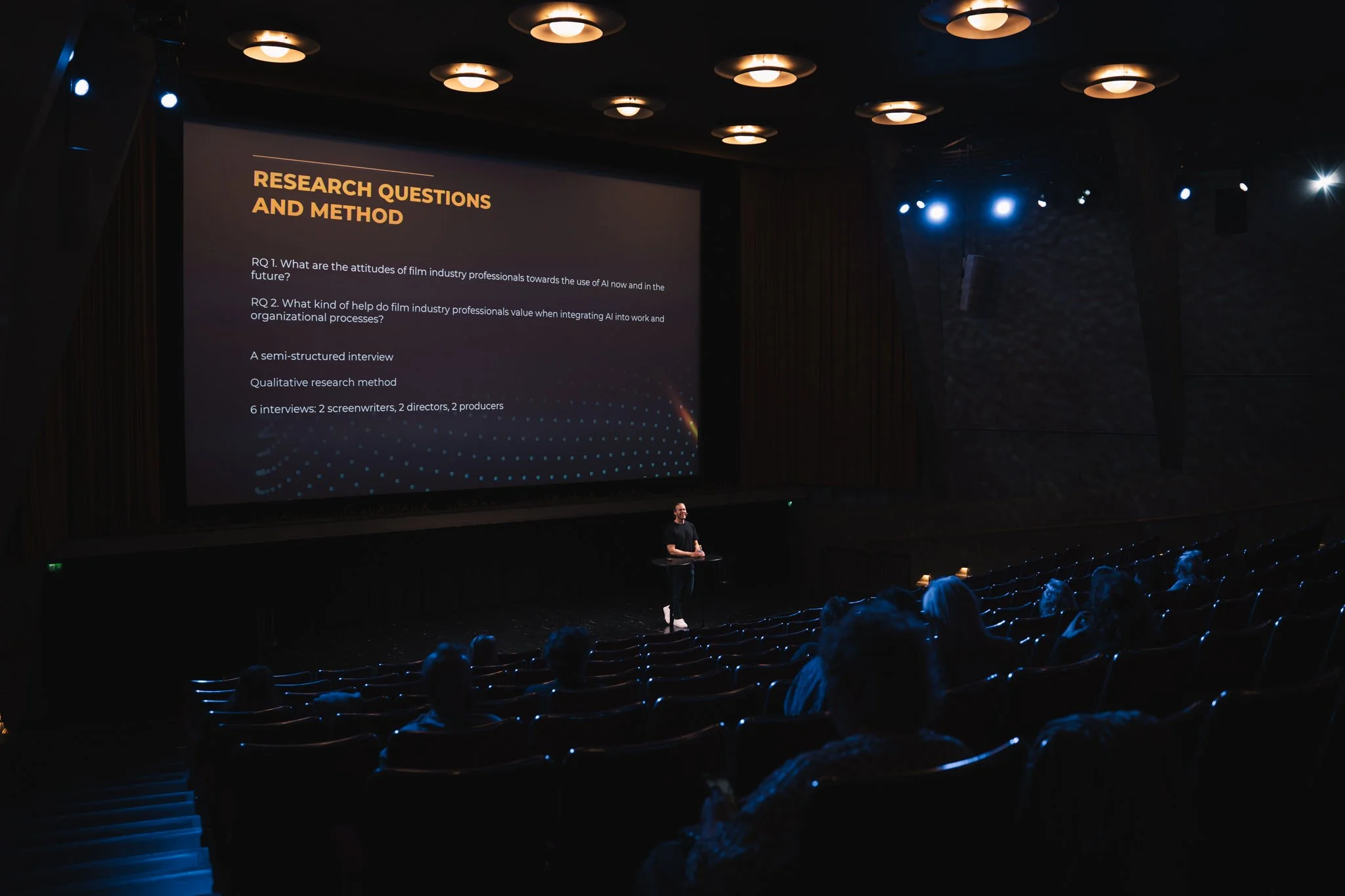 A person presenting on stage in a dark auditorium, illuminated by blue lights, with a large screen displaying research questions and methodology.