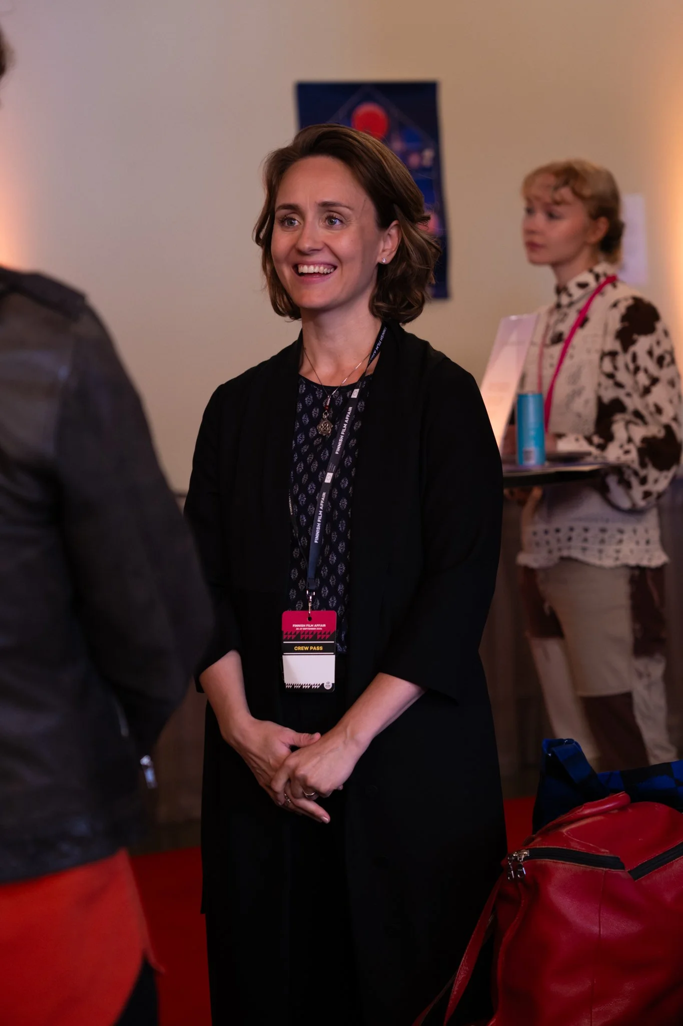 A woman with short brown hair smiling and wearing a black jacket and conference badge is speaking with someone. In the background, another woman with curly blonde hair holding a clipboard or tablet, stands near a table with a blue item on it. The set