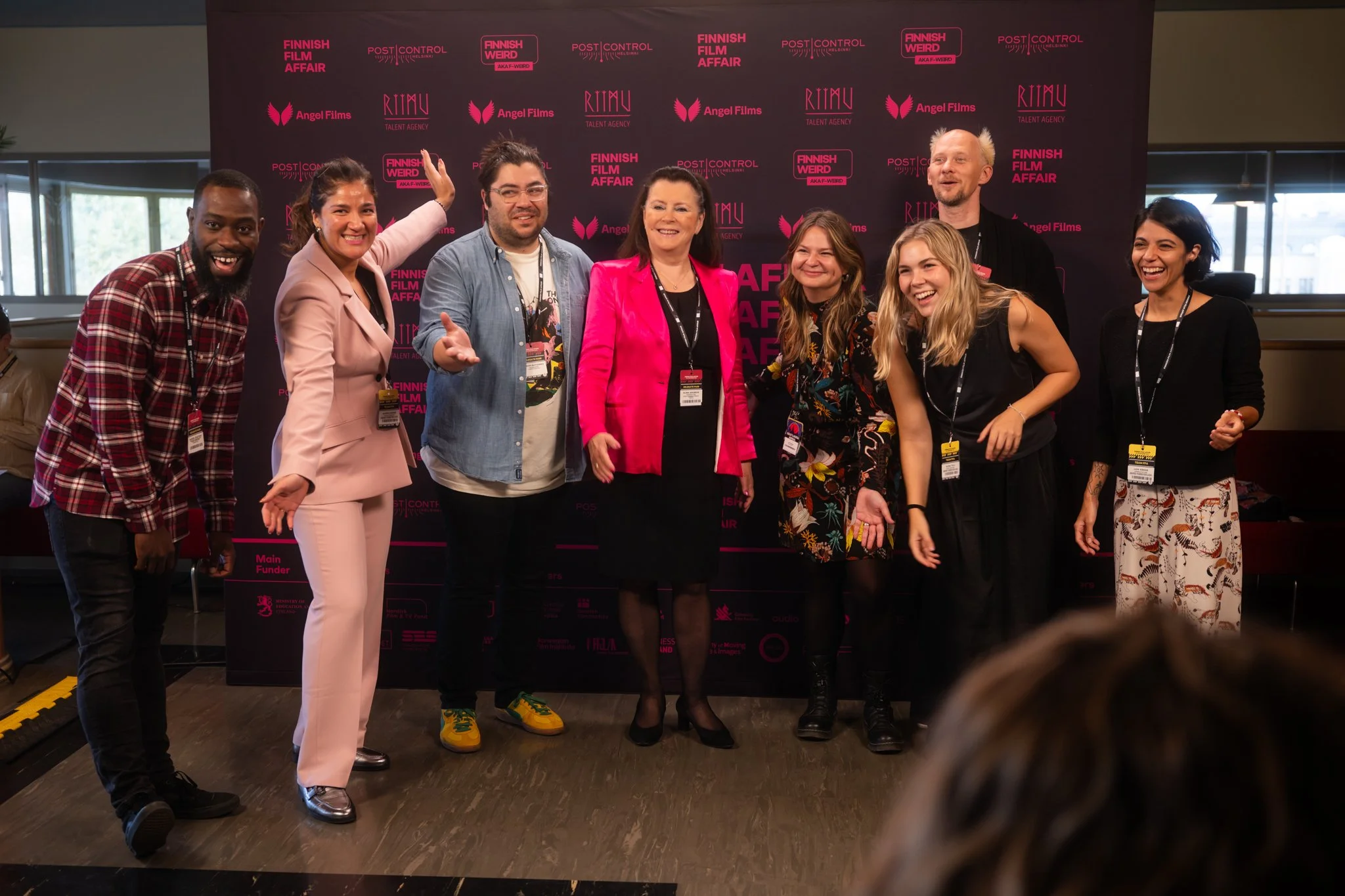 Group of eight people standing in front of a dark pink backdrop with event logos and text, smiling and posing for a photo at the Finnish Film Affair event.