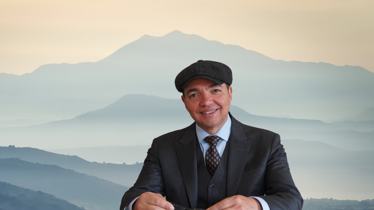 A man in a suit and tie with a flat cap smiling in front of a mountain landscape with layers of mountains in the background.