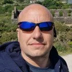 Man wearing sunglasses outdoors on a sunny day, with greenery and stone steps in the background.