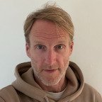 Portrait of a middle-aged man with light hair and beard, wearing a beige hoodie, looking directly at the camera.
