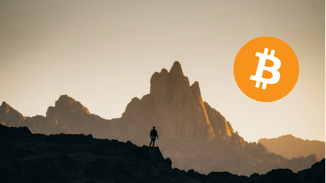 A person standing on rocky terrain in front of mountain peaks during sunset or sunrise, with a large Bitcoin logo overlaid in the sky.