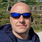 Close-up of a man with a shaved head wearing dark sunglasses and a dark jacket, outdoors with greenery and stone steps in the background.