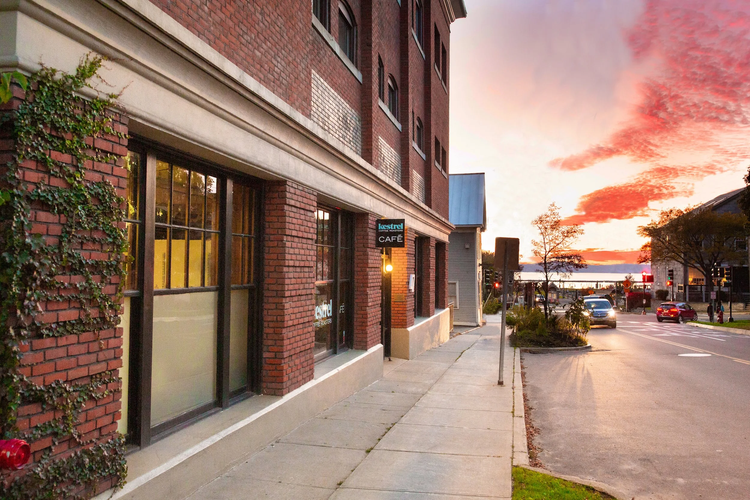 Front of Karma Bird House, a brick building with a sign for Kestrel cafe, sidewalk, street with cars, and a colorful sunset sky with clouds and water in the distance.