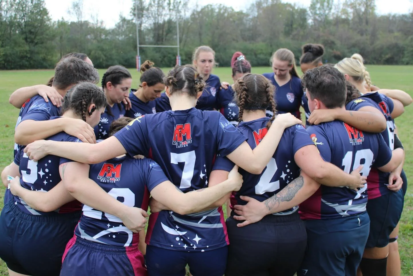A team of women rugby players in blue jerseys huddles on a grass field with a goalpost and trees in the background.
