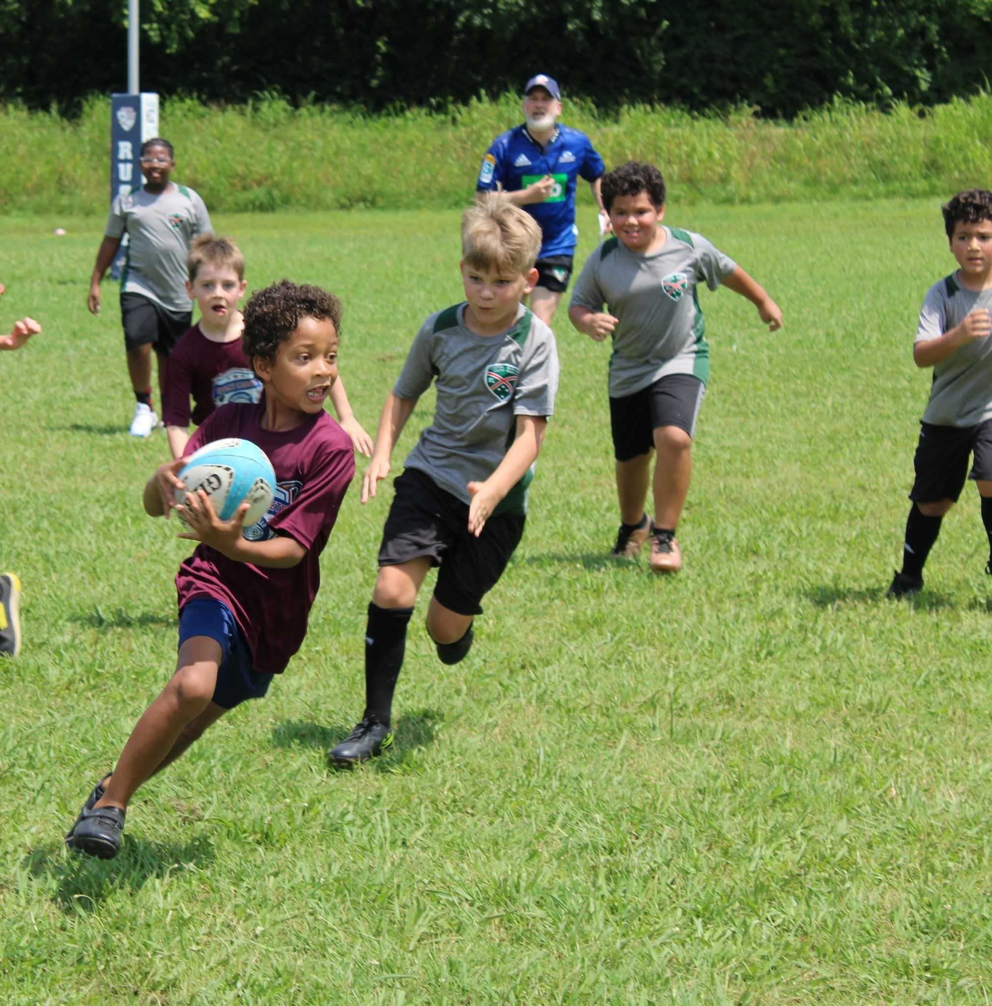 Children playing rugby on a grassy field, with one child running with the ball and adults and other kids chasing behind.
