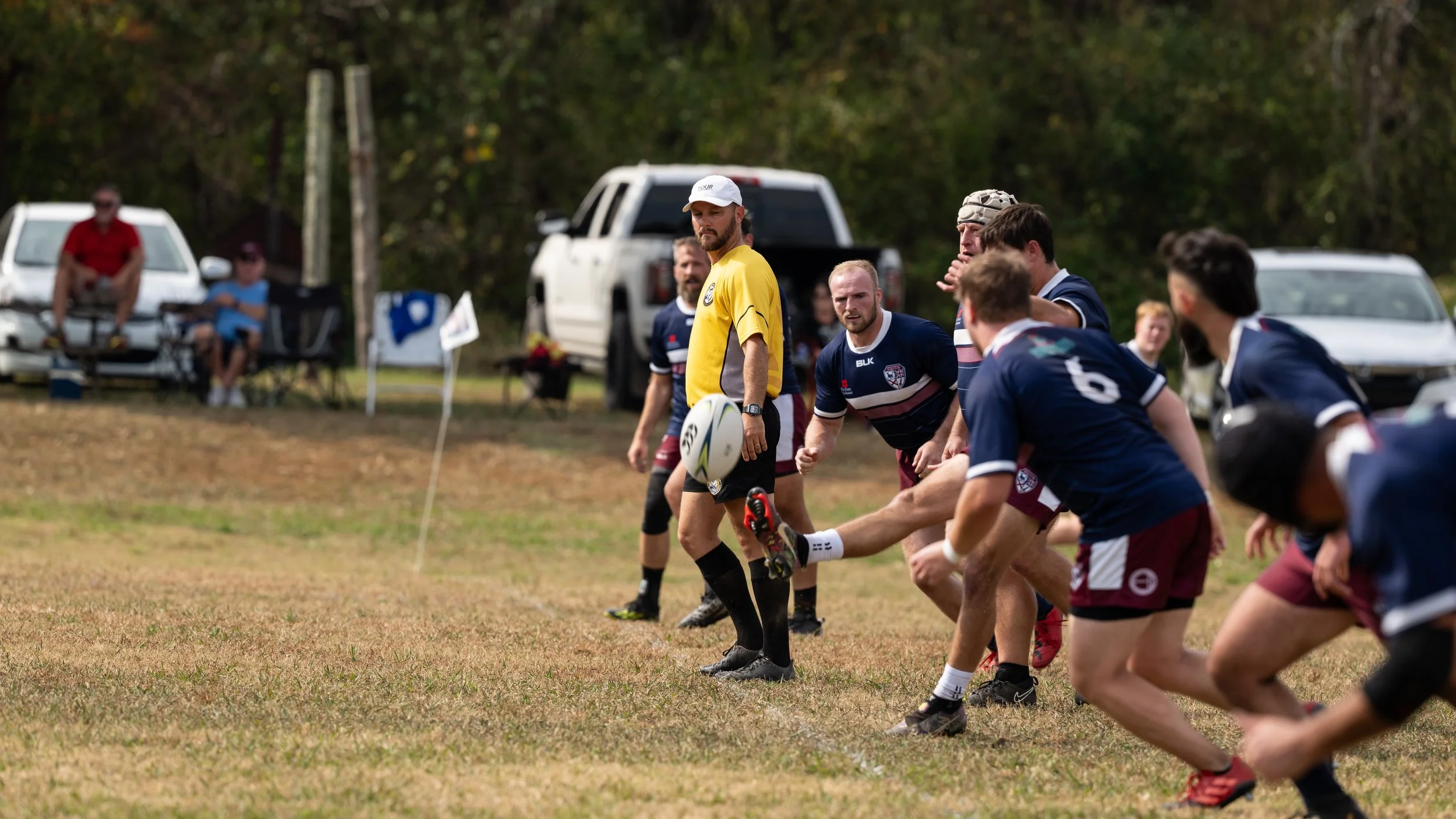 A rugby match in progress with players in navy and maroon uniforms, one player kicking the ball, and a referee in a yellow shirt standing nearby on a grassy field. Spectators and vehicles are visible in the background.