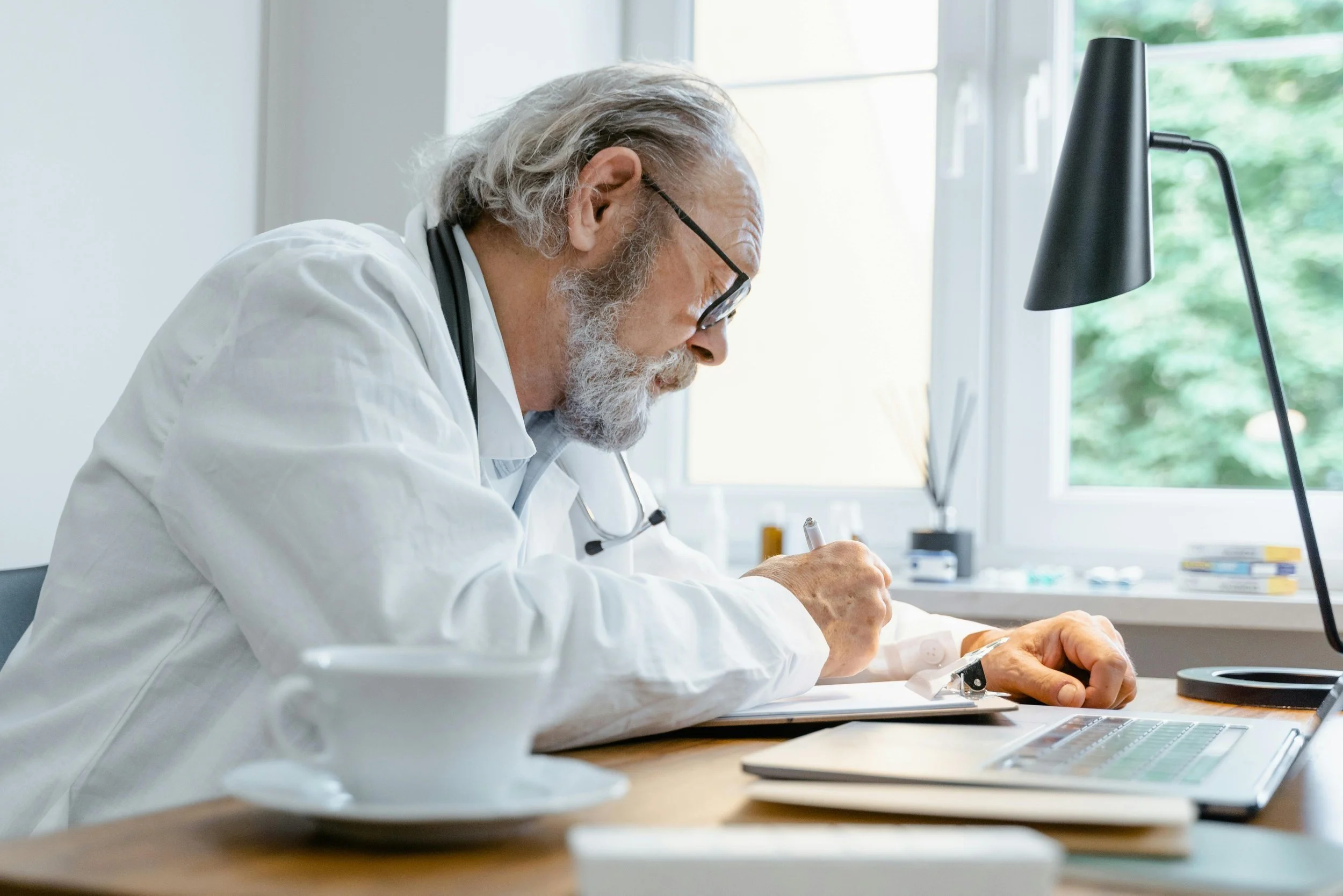 An elderly male doctor with gray hair, beard, and glasses writing on a clipboard at a desk in a medical office, with a coffee cup, laptop, and window in the background.