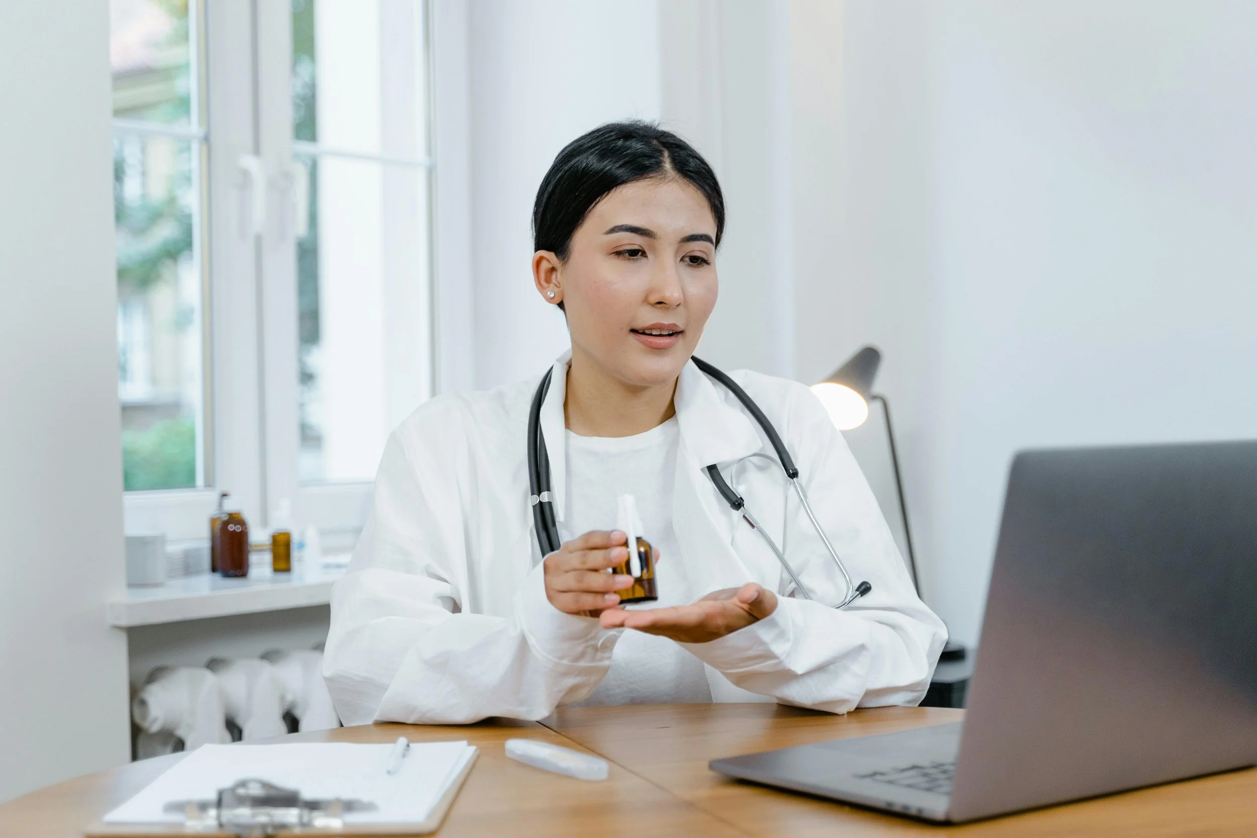 A female doctor in a white coat with a stethoscope around her neck sitting at a desk, holding a small medicine bottle and looking at a laptop, with medicine bottles on a shelf behind her.