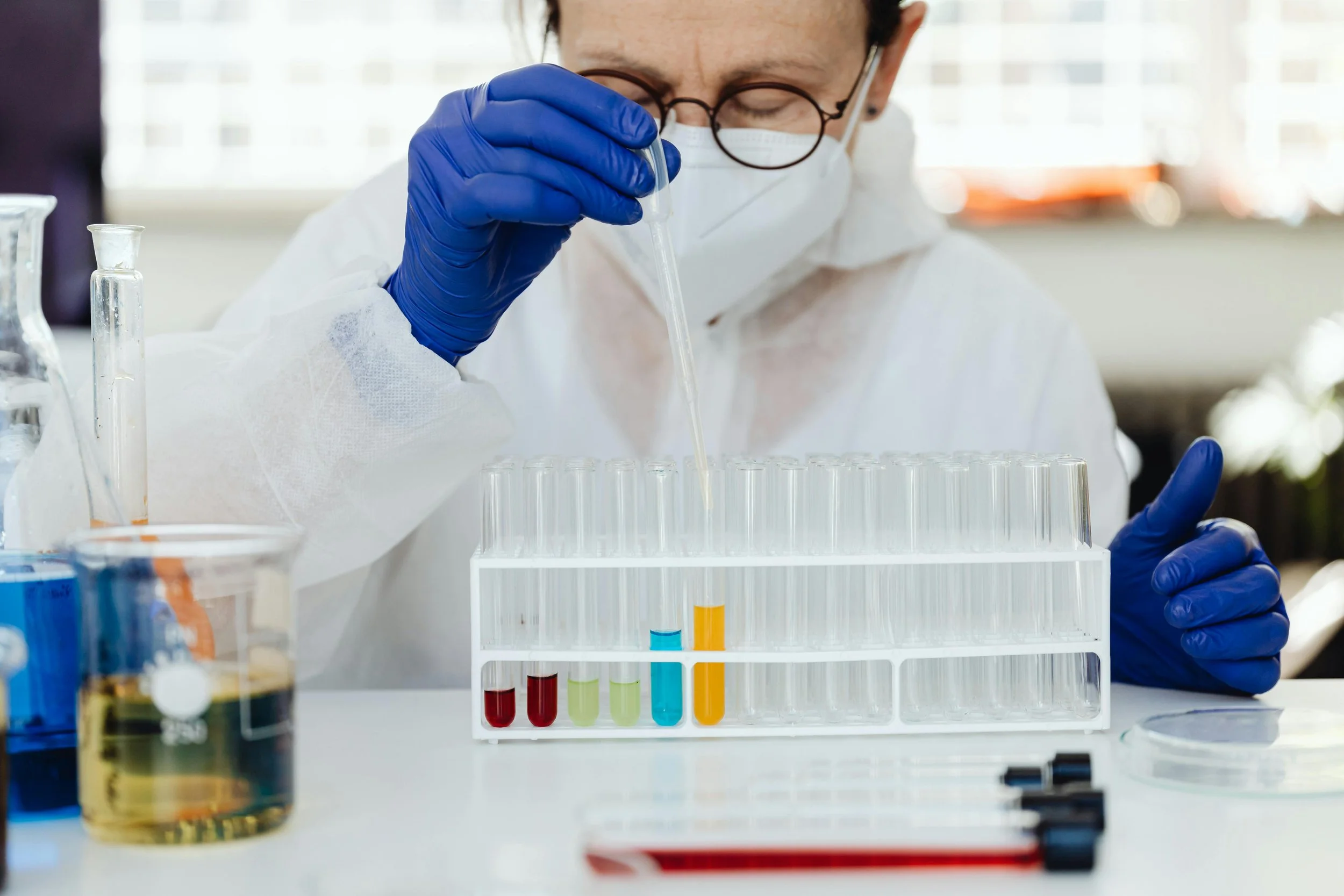 A scientist wearing a mask, glasses, and blue gloves using a pipette to transfer liquid into test tubes in a laboratory.