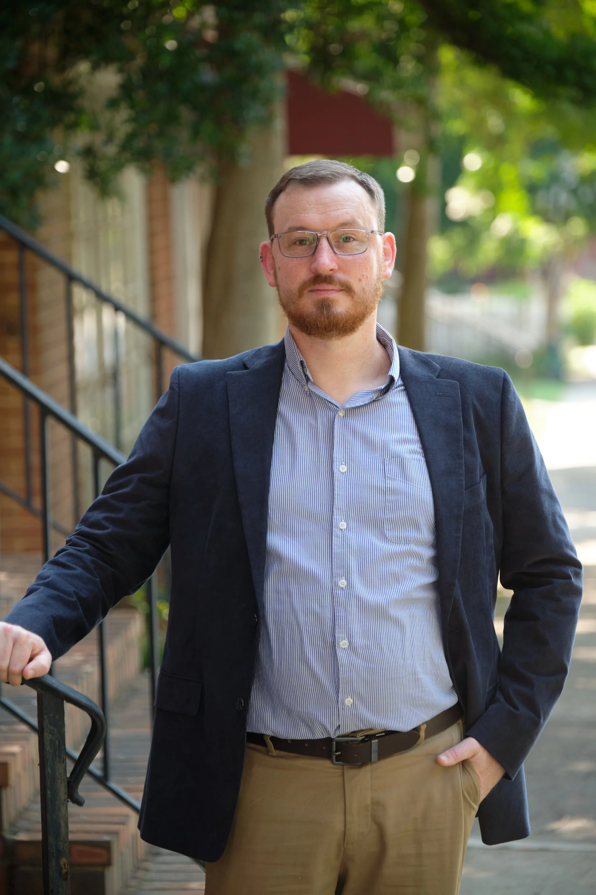 A man with glasses, a beard, and wearing a navy blazer and light blue shirt stands outdoors on a sunny day, with trees and a sidewalk in the background.