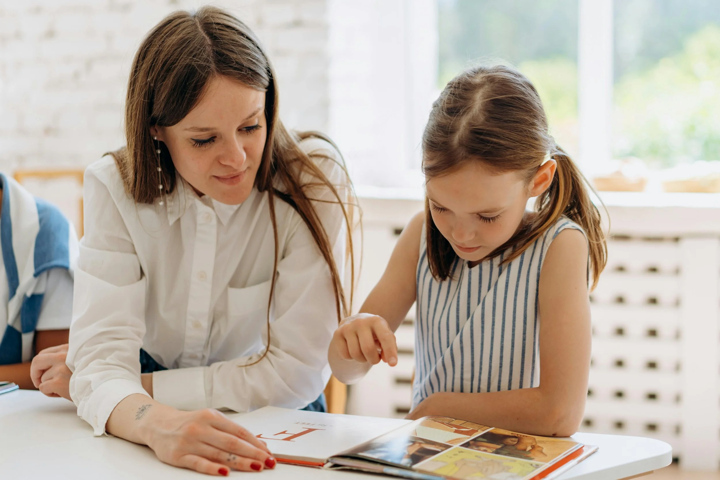 A woman and a young girl looking at a colorful book together at a table in a bright room.