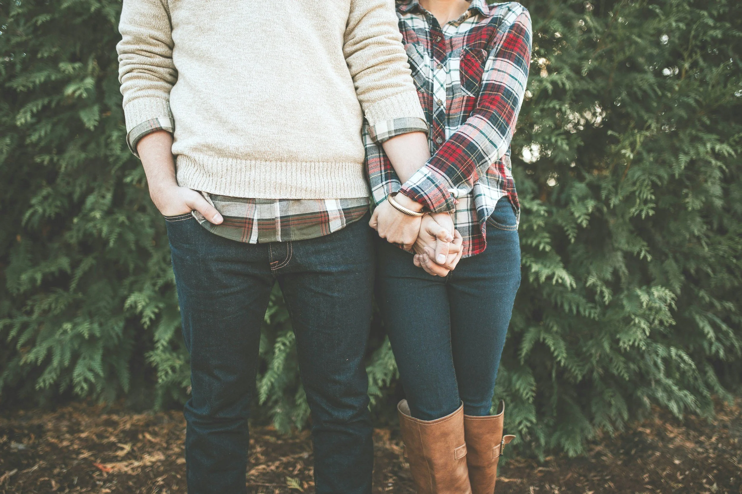 A couple holding hands in front of greenery, wearing casual fall clothing.