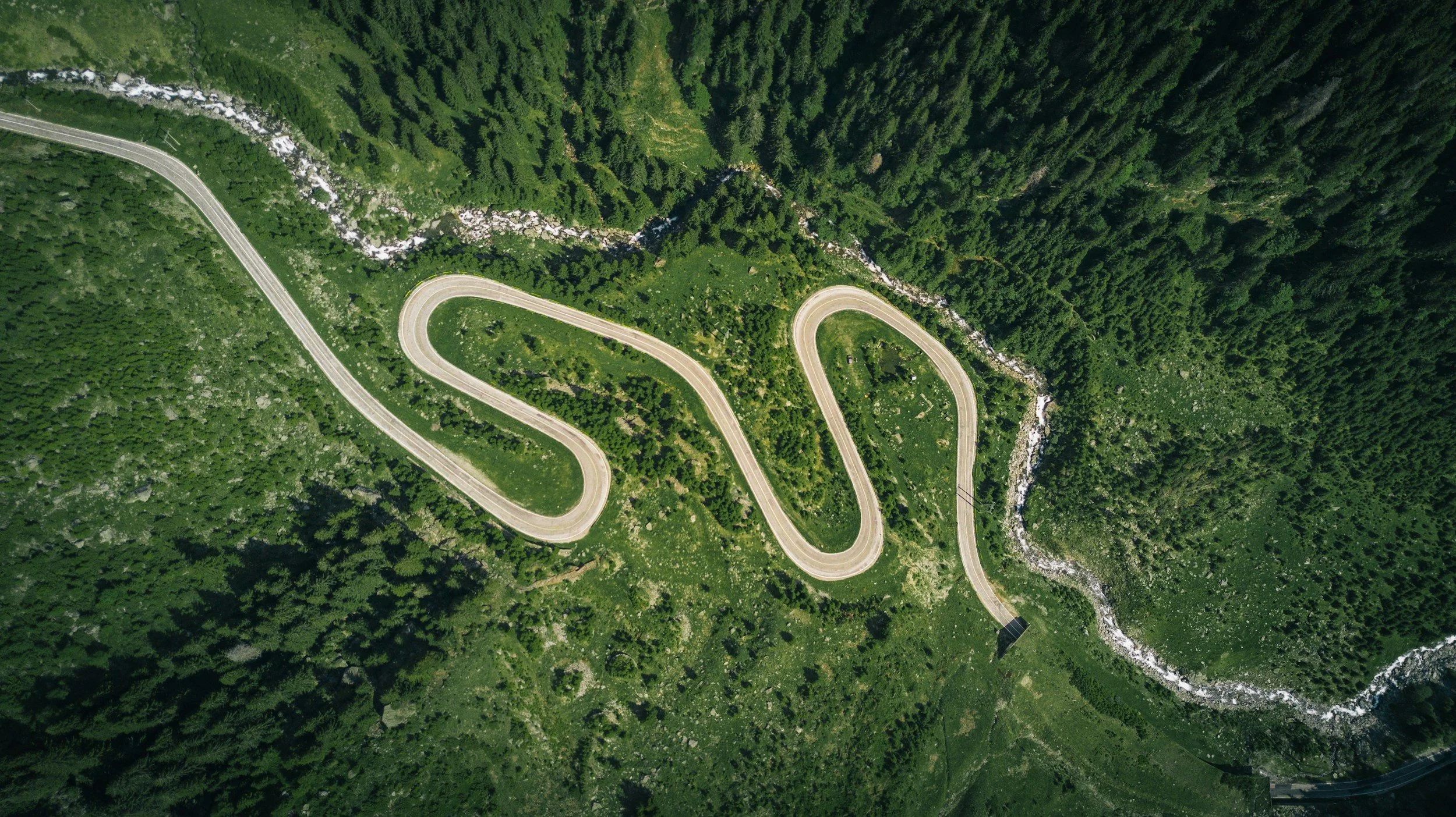 An aerial view of a winding mountain road through a forested area with lush green trees and rocky terrain.