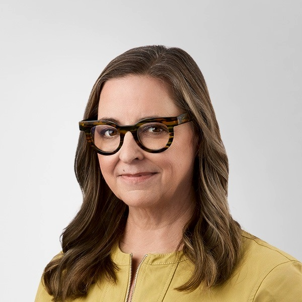 A woman with long brown hair wearing large tortoise shell glasses and a yellow jacket, smiling softly against a plain background.
