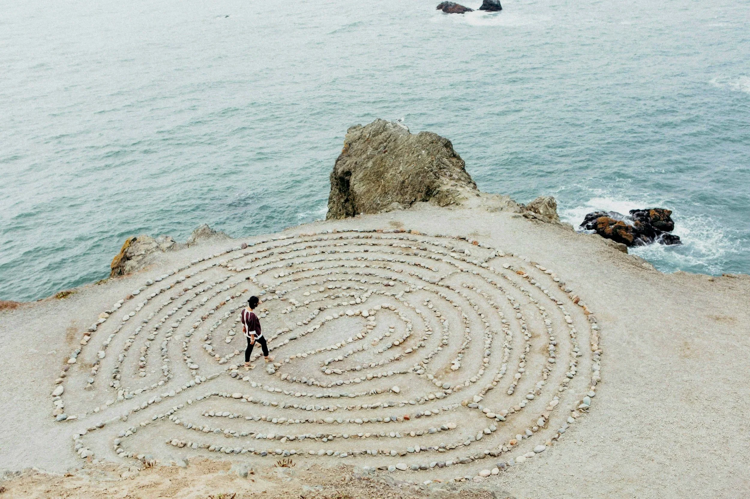 A person walking in a spiral pattern made of rocks on a sandy area near the ocean, with rocks and water in the background.