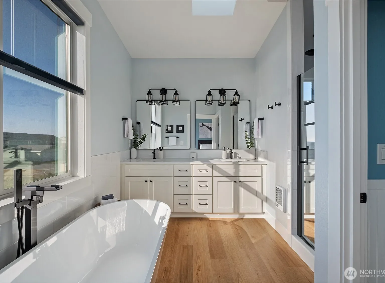 Modern bathroom with white vanity, dual sinks, large mirror, black fixtures, wood flooring, bathtub, and a glass-enclosed shower, with natural light coming through a window.