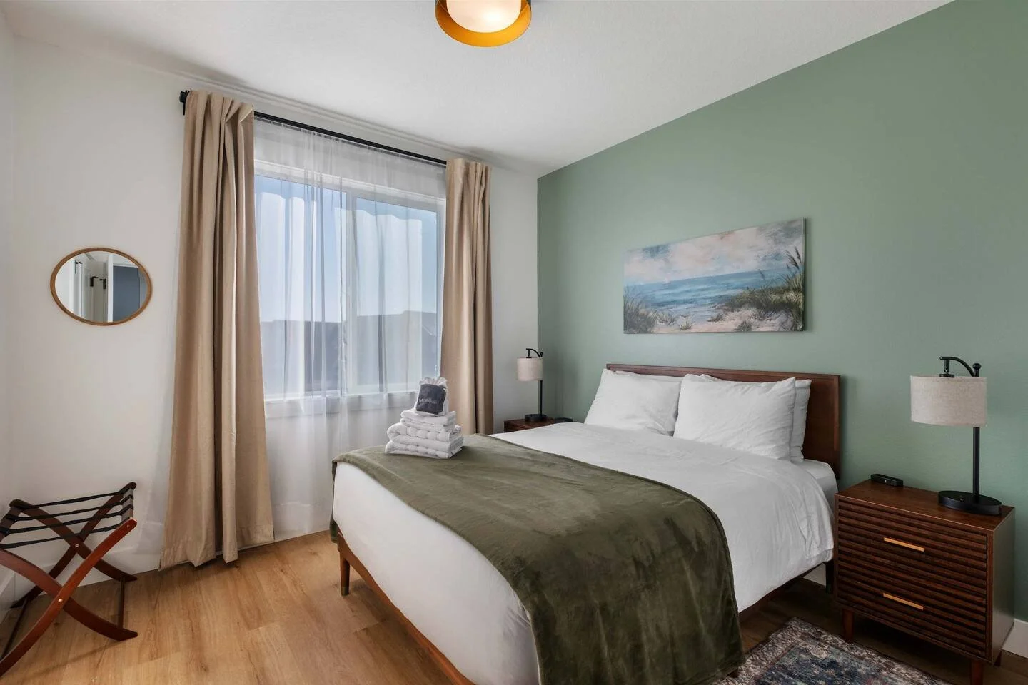 A modern bedroom with a bed, green accent wall, ocean-themed artwork, two nightstands with lamps, and a window with beige curtains.