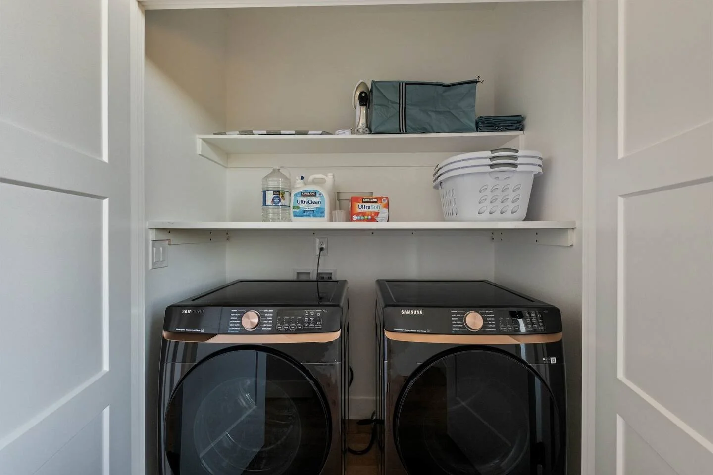 Laundry room with a black Samsung washer and dryer, white shelves above containing cleaning supplies, a laundry basket, and folded clothes.