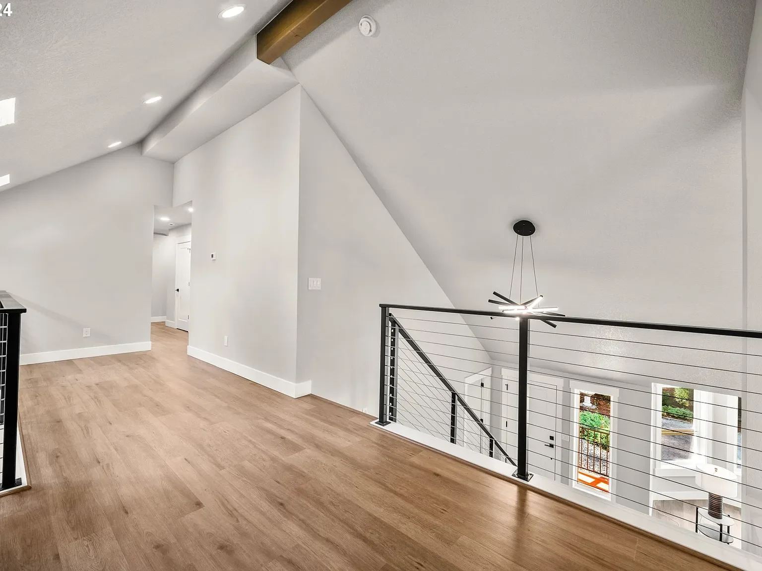 Interior view of a modern, bright home with white walls, wooden floors, a black railing, a ceiling fan with lights, and large windows.
