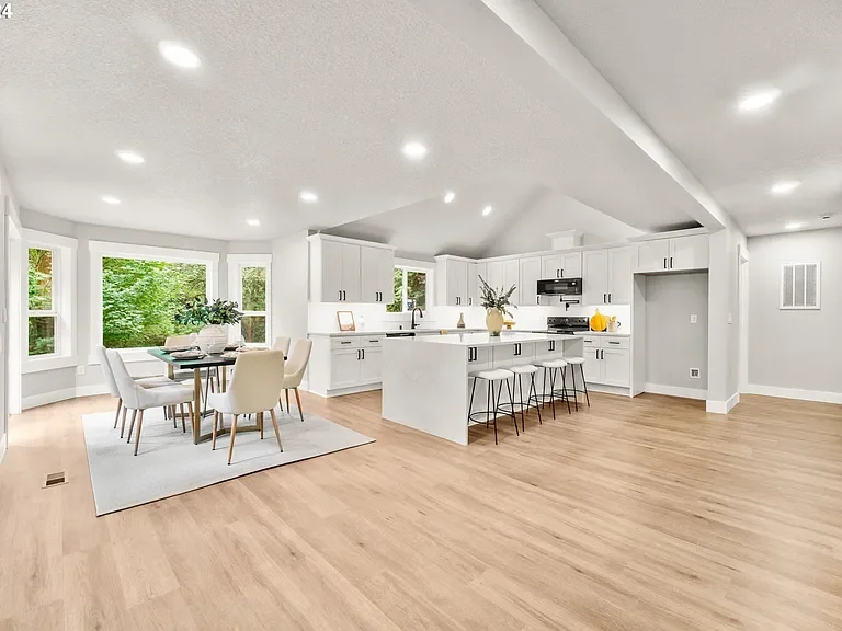 Open-concept kitchen and dining area with white cabinets, a kitchen island, and large windows overlooking greenery.