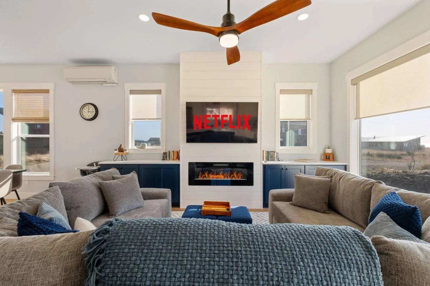 Living room with beige sofas, blue pillows, a fireplace, a wall-mounted TV displaying the Netflix logo, windows with blinds, large windows showing an outdoor view, and a ceiling fan.