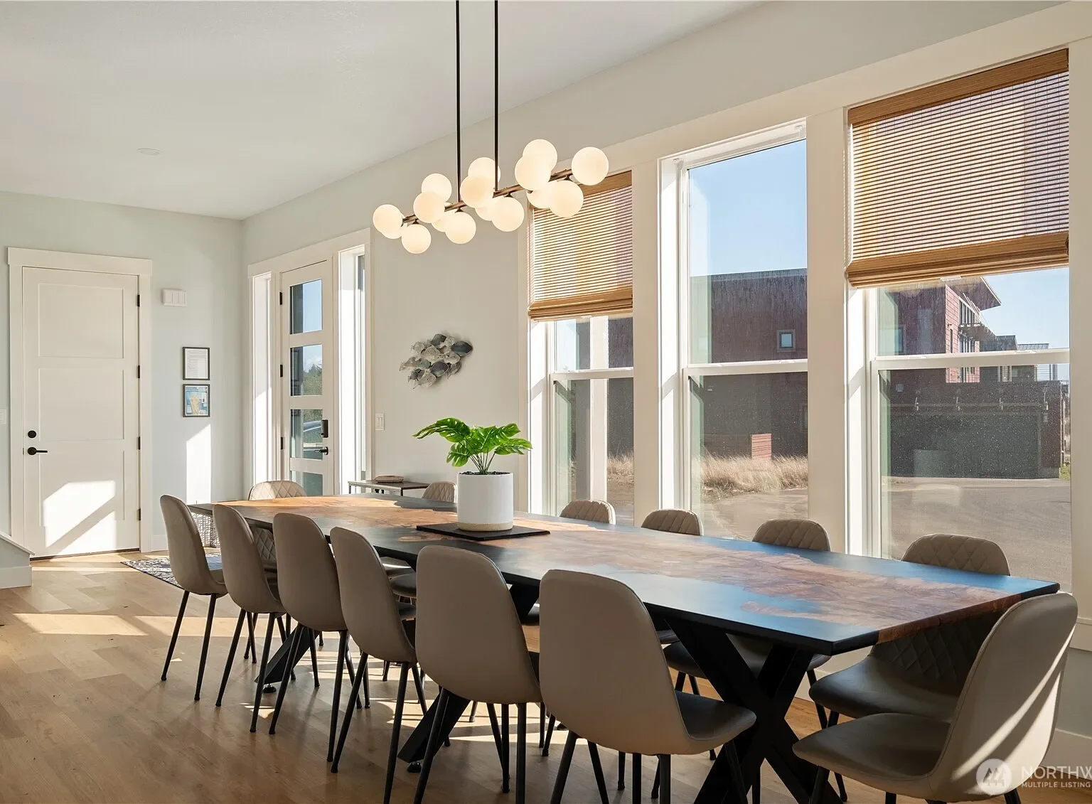 Modern dining room with a large wooden table surrounded by beige chairs, a plant centerpiece, large windows with wooden blinds, and a contemporary chandelier with white globe lights.