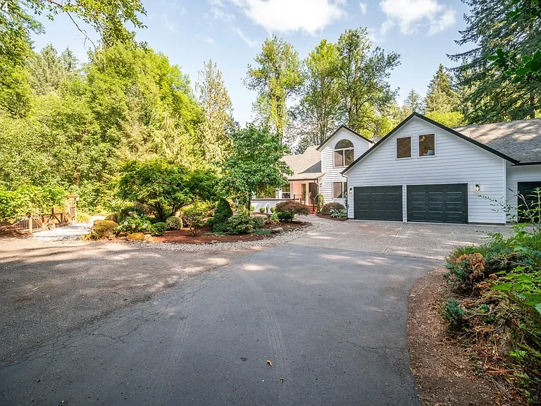 A large house with a white garage and dark doors, surrounded by greenery and trees, with a curved driveway in front.