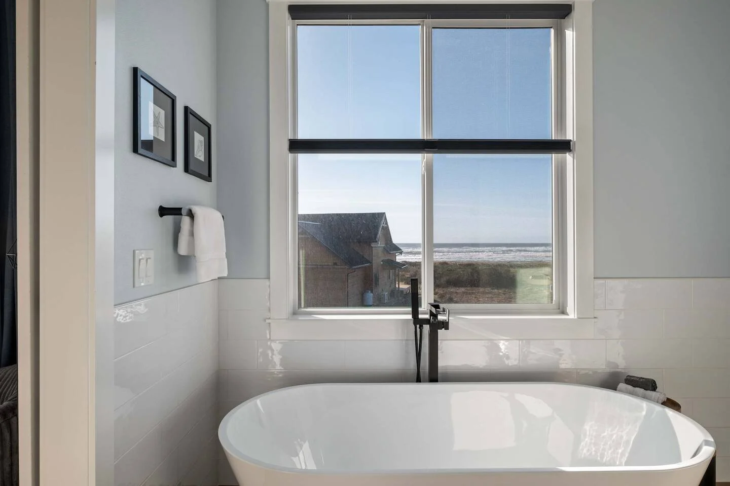 Modern bathroom with a large window overlooking a beach and houses, a white bathtub, black faucet, and framed wall art.