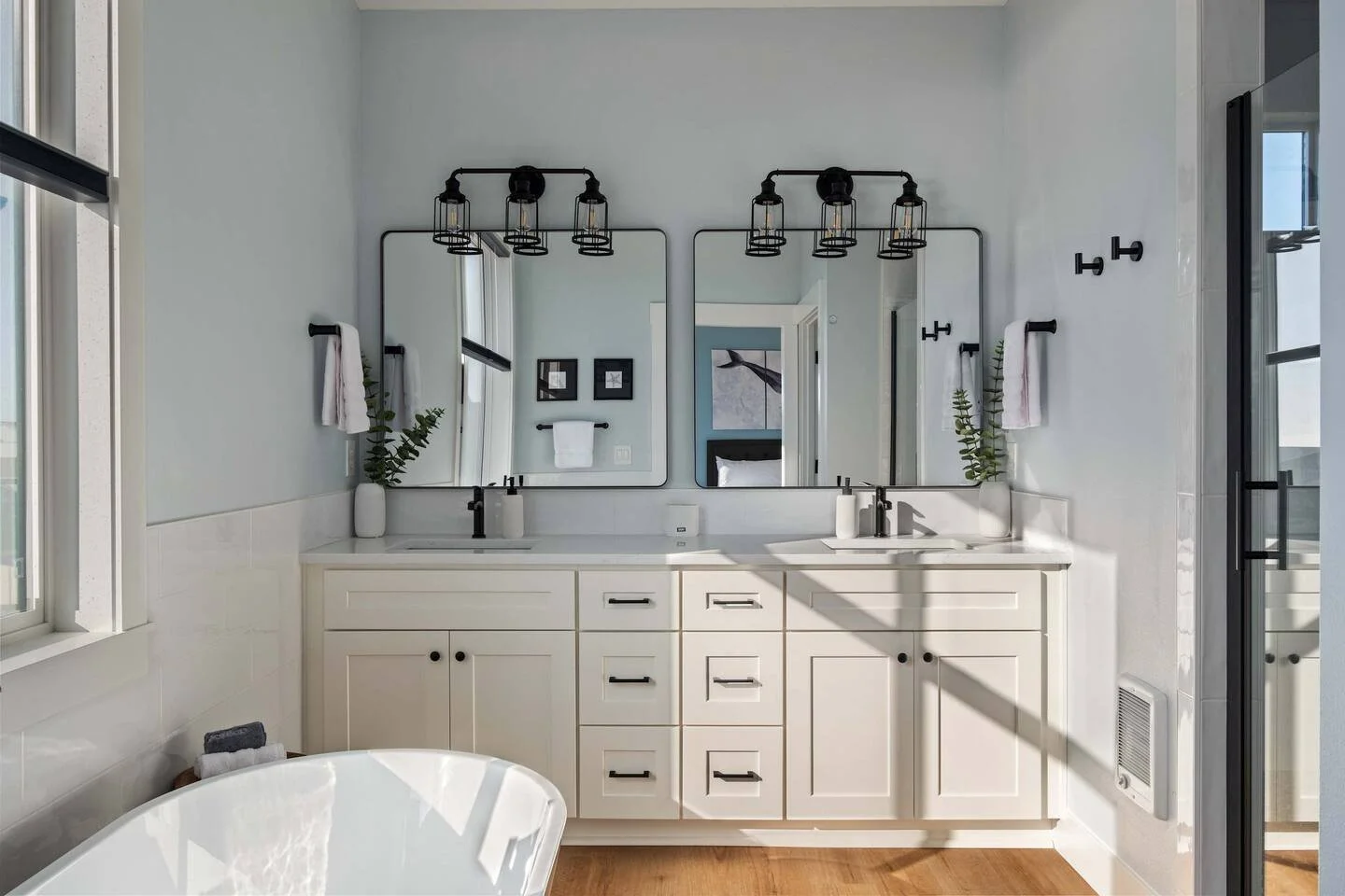 Modern bathroom with dual sinks, large mirrors, black light fixtures, white cabinetry, and a clawfoot bathtub. Two small plants on the countertop and towels hanging on black hooks.