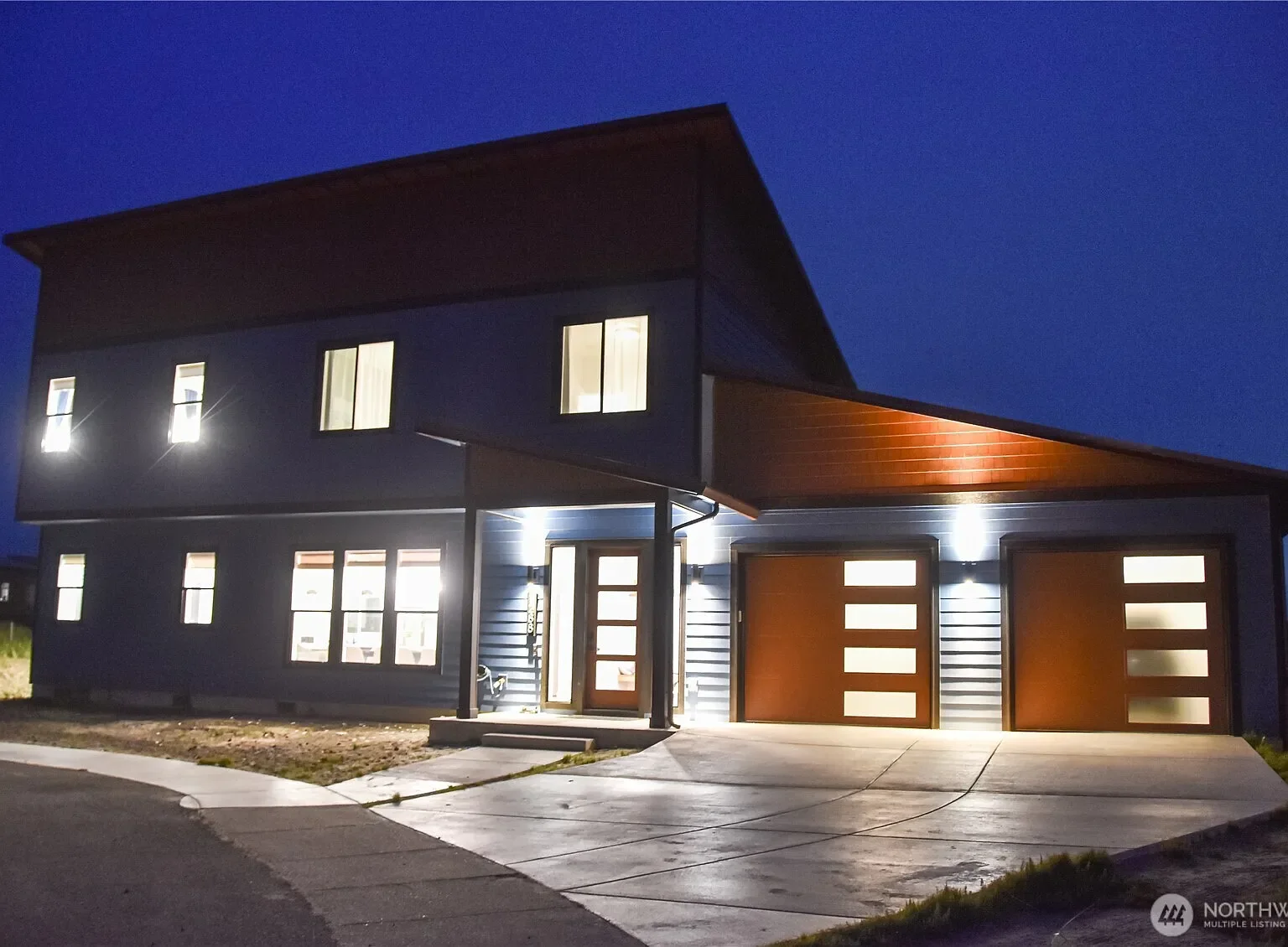 Modern two-story house with blue siding and wooden garage doors illuminated at night.