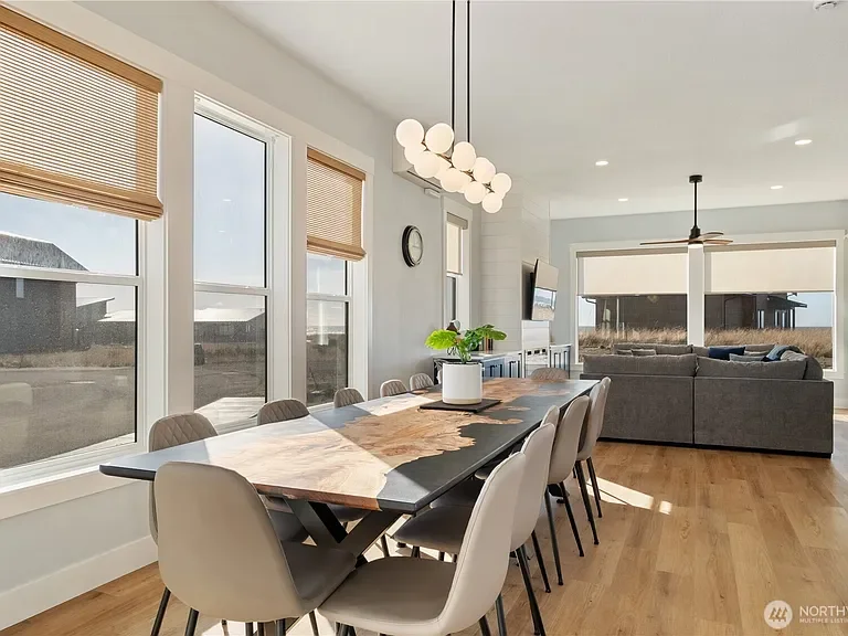 Bright dining area with large windows, a wooden table, and multiple chairs, connected to a living room with a gray sectional sofa and a TV.