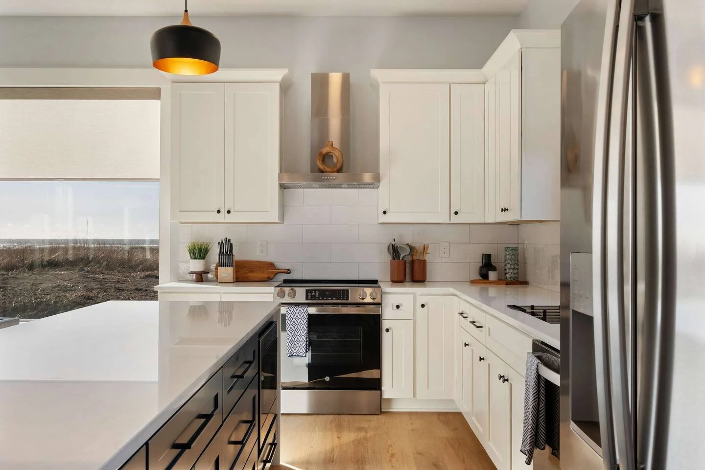 Modern kitchen with white cabinets, stainless steel appliances, a black and gold pendant light, and a view of the outdoors through a large window.