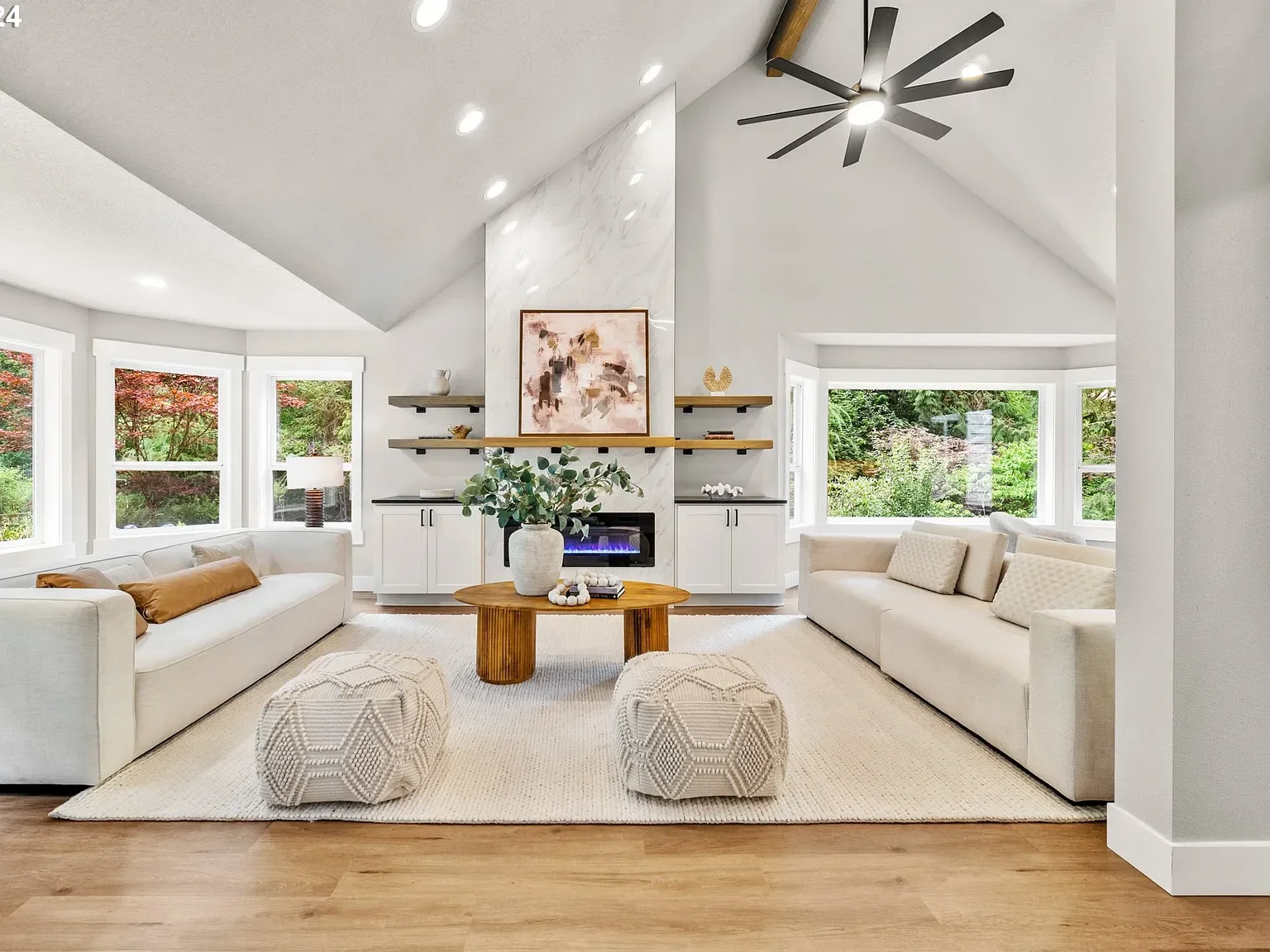 A bright living room with a vaulted ceiling, large windows, two white sofas, two textured ottomans, a round wooden coffee table with a vase of greenery, and a fireplace with abstract artwork above and floating shelves on each side.