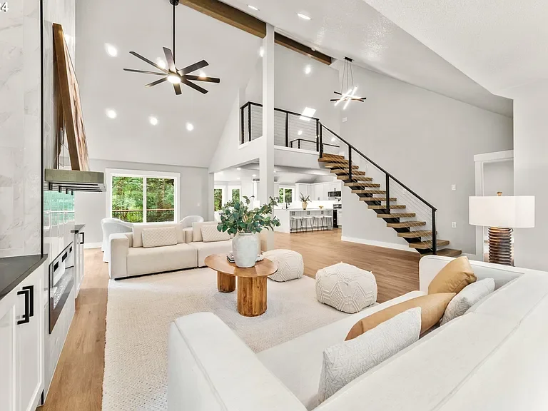 Open-concept living room with high vaulted ceiling, white sofas, a wooden coffee table with greenery, ottomans, and modern lighting fixtures.