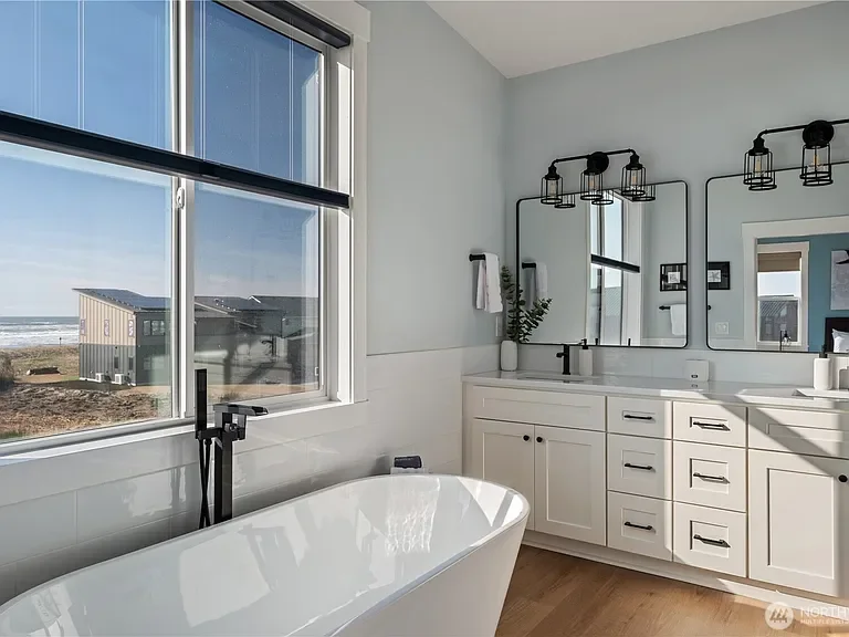 Bathroom with a freestanding bathtub, large window showing a beach view, white vanity with dual mirrors, and black fixtures.