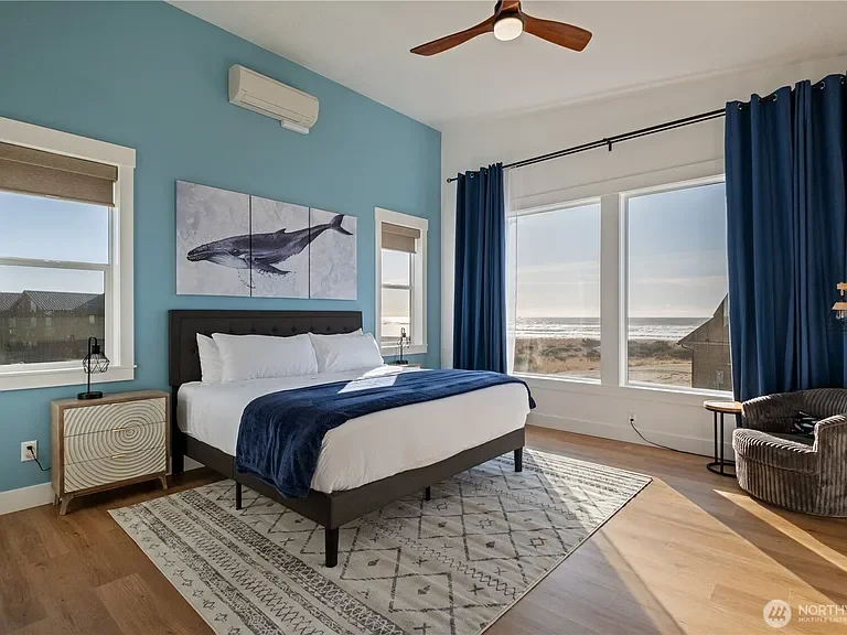 Bedroom with light blue walls, large windows, and dark blue curtains. A bed with white bedding and a dark blue blanket is centered against the wall, with a whale art piece hanging above. A wooden side table with a lamp is next to the bed. There is a