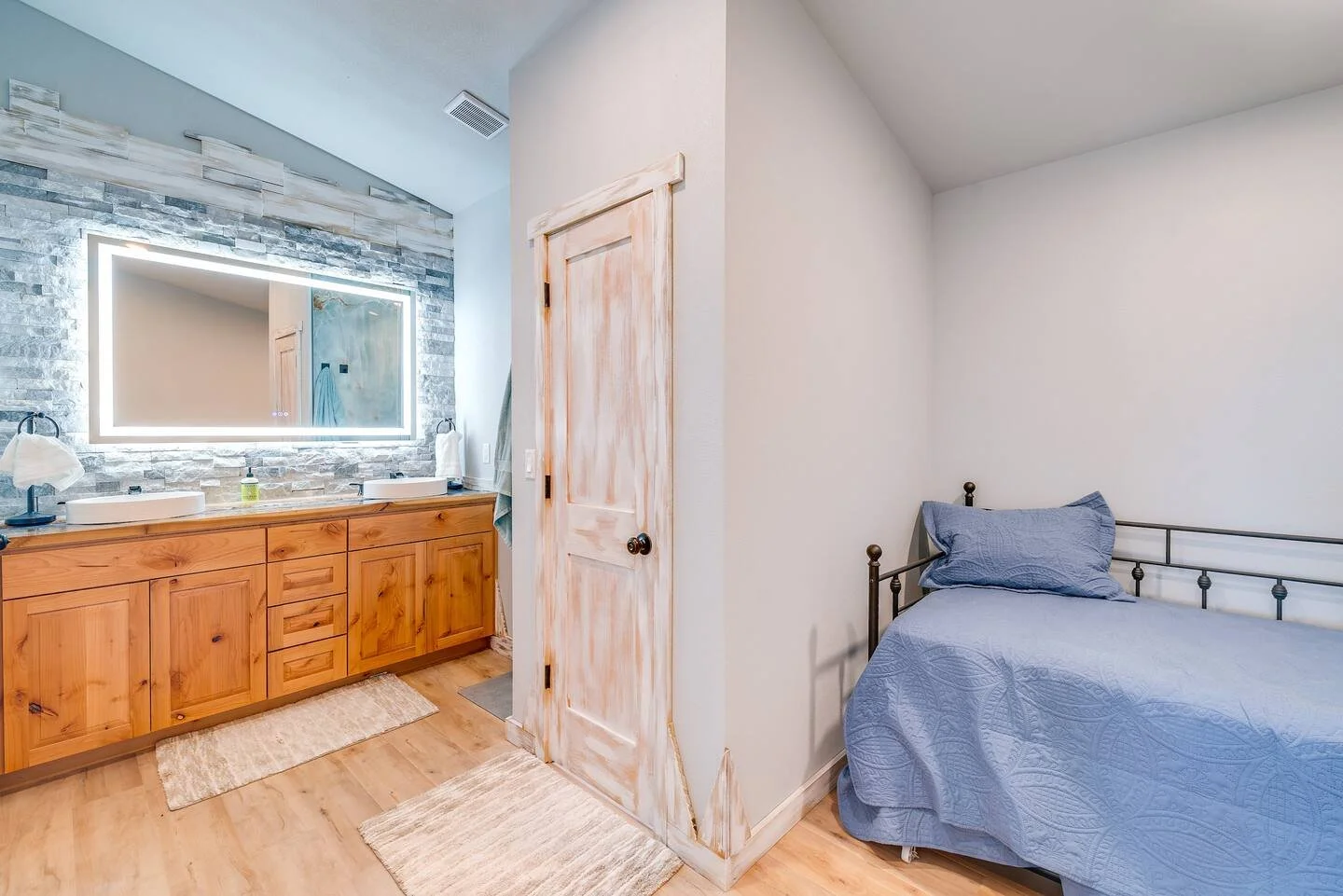 Bedroom with a wooden dresser and large mirror in the bathroom, and a metal bed with blue bedding in the corner.