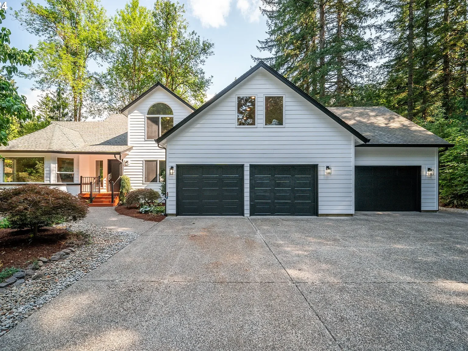 A modern white house with black garage doors, surrounded by trees and a well-maintained yard, featuring a driveway and a small front porch.