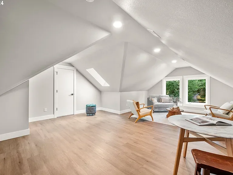 Empty attic room with sloped ceiling, hardwood floors, and white walls, furnished with a small seating area including a gray sofa, armchair, coffee table, and large window with trees outside.