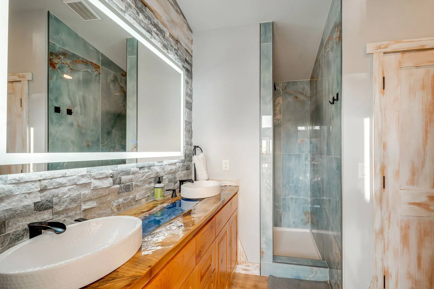 Modern bathroom with dual vessel sinks on wooden countertop, large backlit mirror, stone accent wall, and glass-enclosed shower with blue and orange marbled tiles.