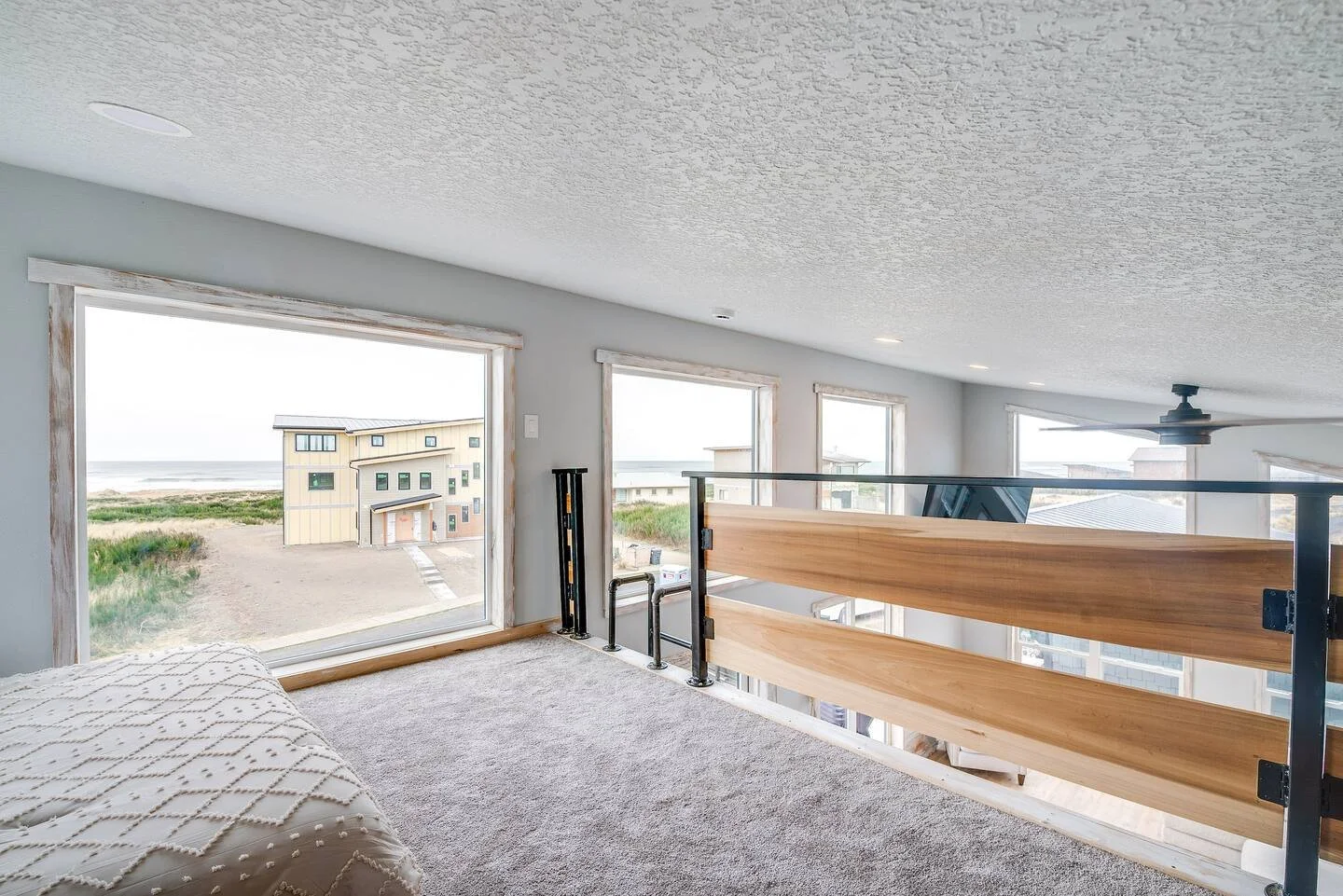 A bright, airy loft with large windows overlooking a beach and neighboring houses, featuring a bed with a textured white blanket, carpeted floor, and a black ceiling fan.