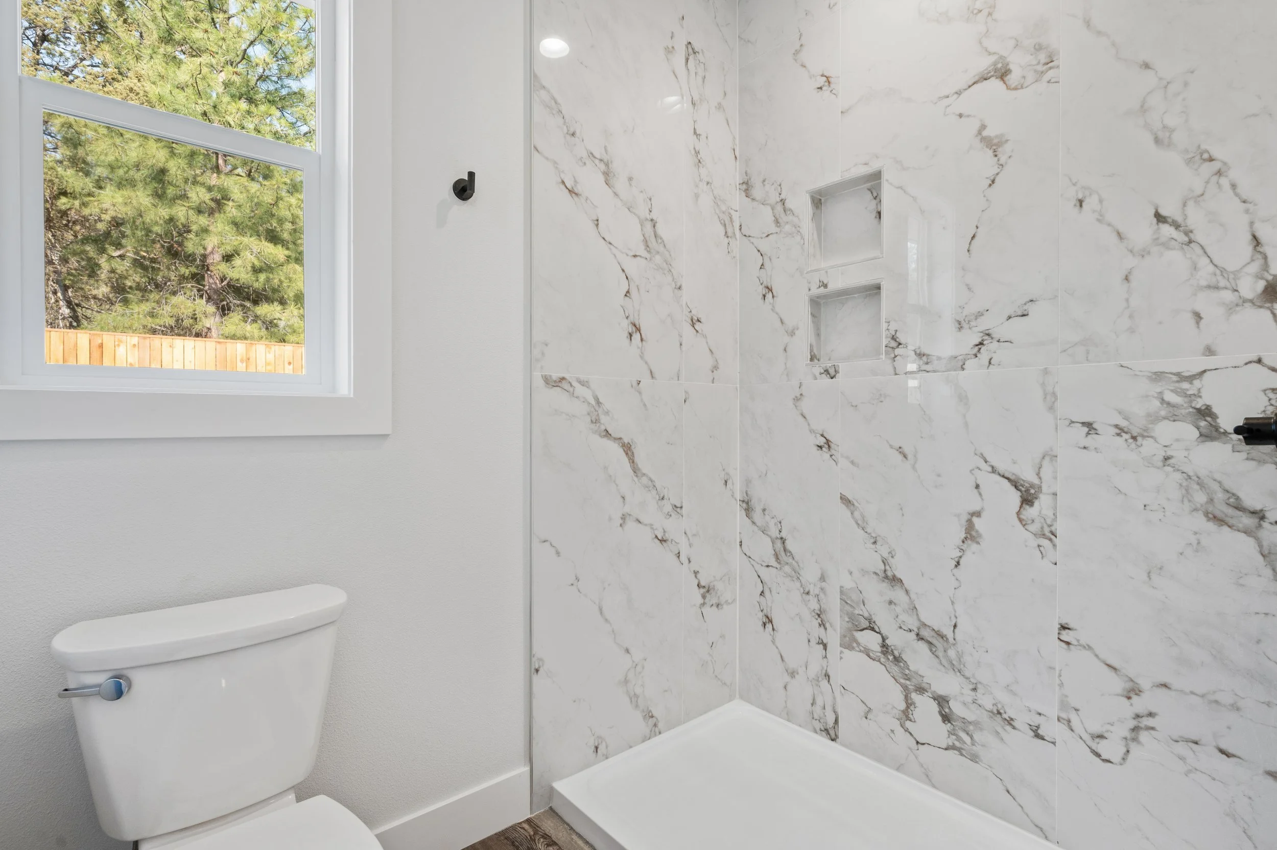 Bathroom with white toilet, marble wall tiles in the shower area, built-in shelves, a window showing outdoor trees, and a small black hook on the wall.