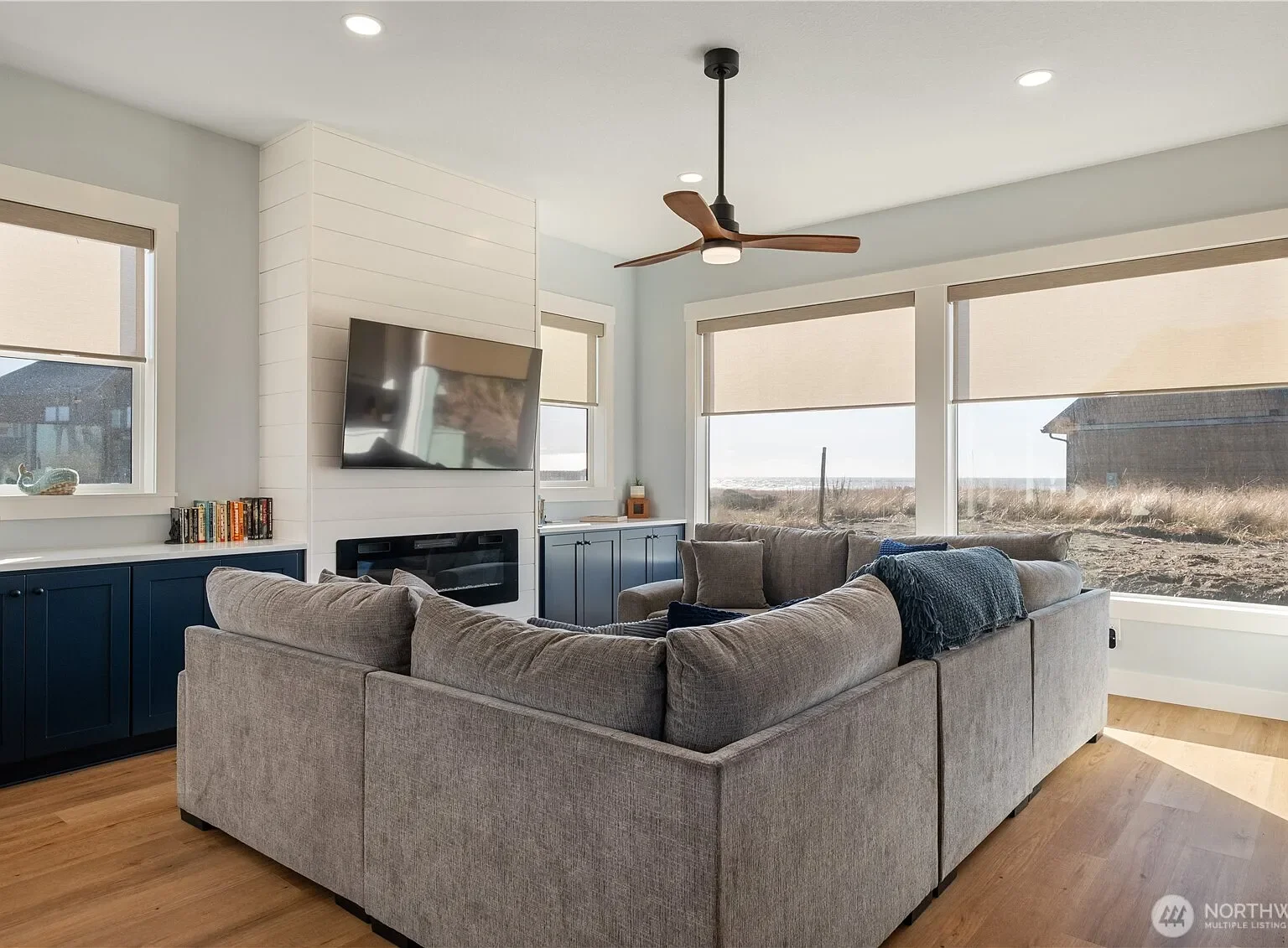 Living room with large windows, beige sectional sofa, ceiling fan, mounted flat-screen TV above a modern fireplace, dark blue cabinetry, and hardwood flooring, with a view of an outdoor landscape.