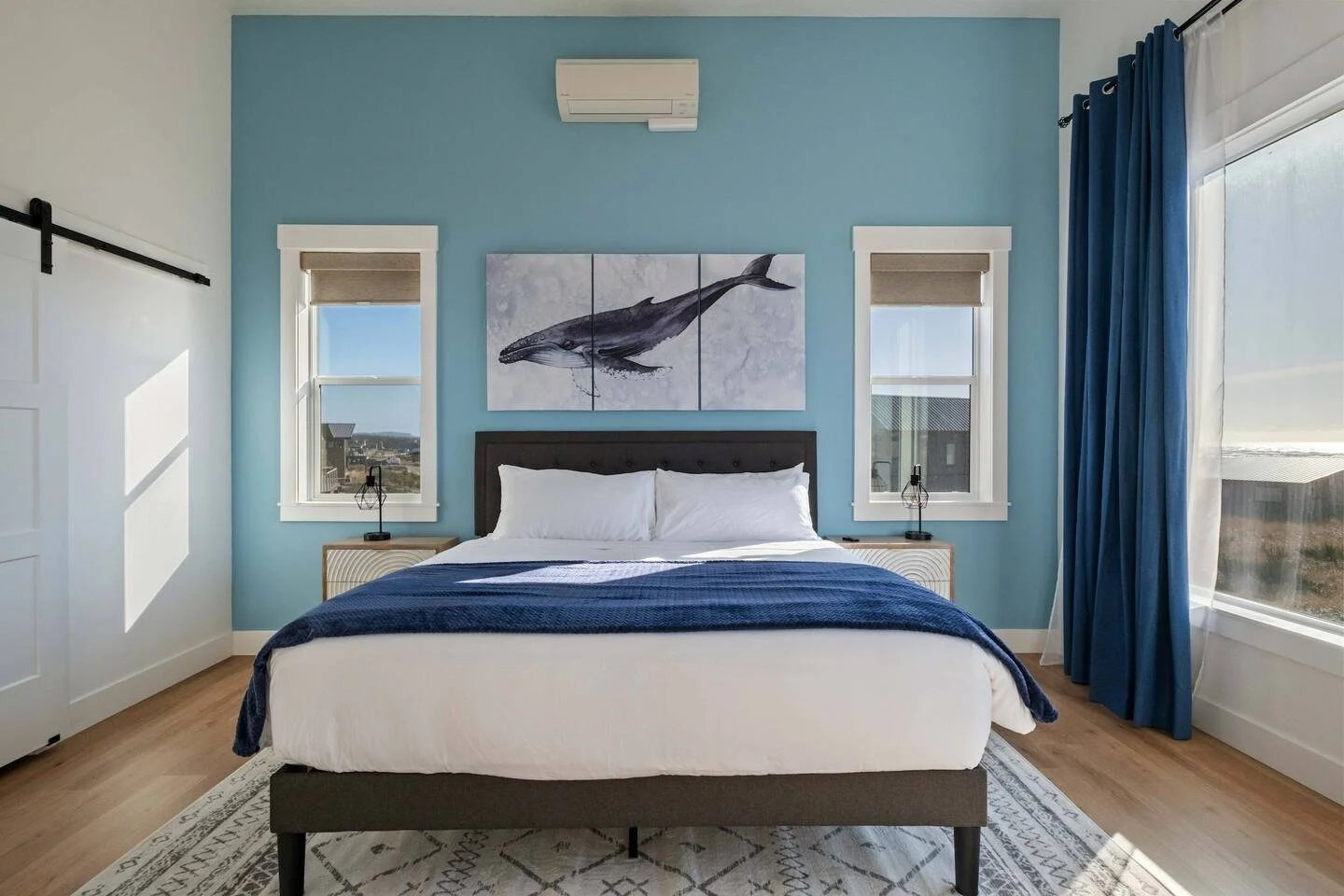 Modern bedroom with a blue accent wall, white bedding, navy throw blanket, and wall art of a whale, illuminated by natural light from large windows with blue curtains.