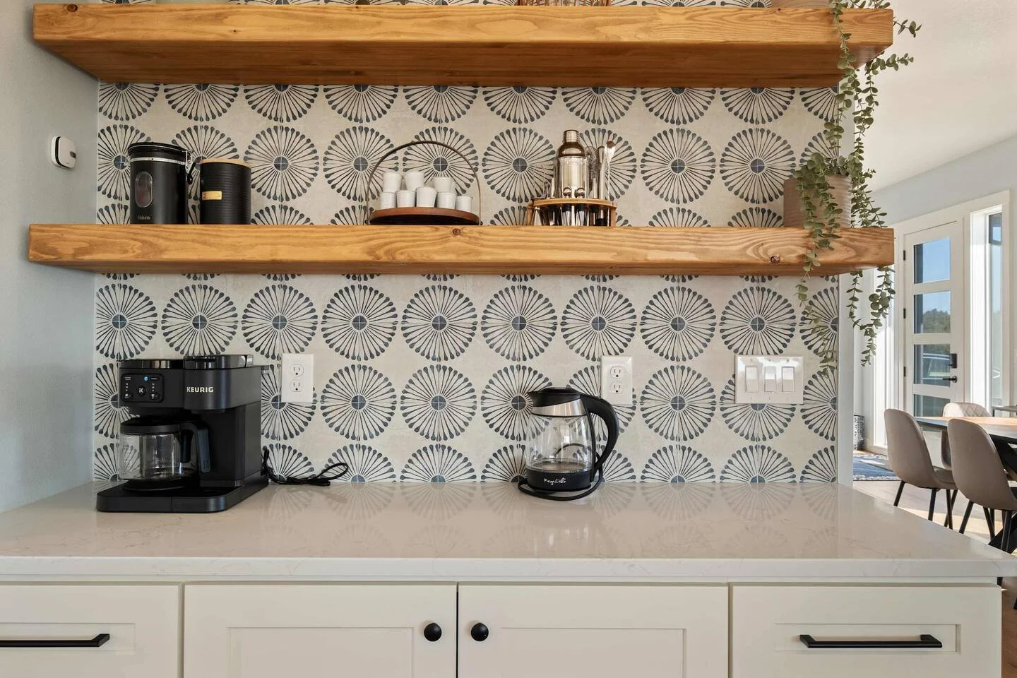 Modern kitchen counter with coffee maker, electric kettle, and glass teapot, and wooden shelves holding cups and drink dispensers. Decor includes patterned wallpaper and a potted vine in the corner.