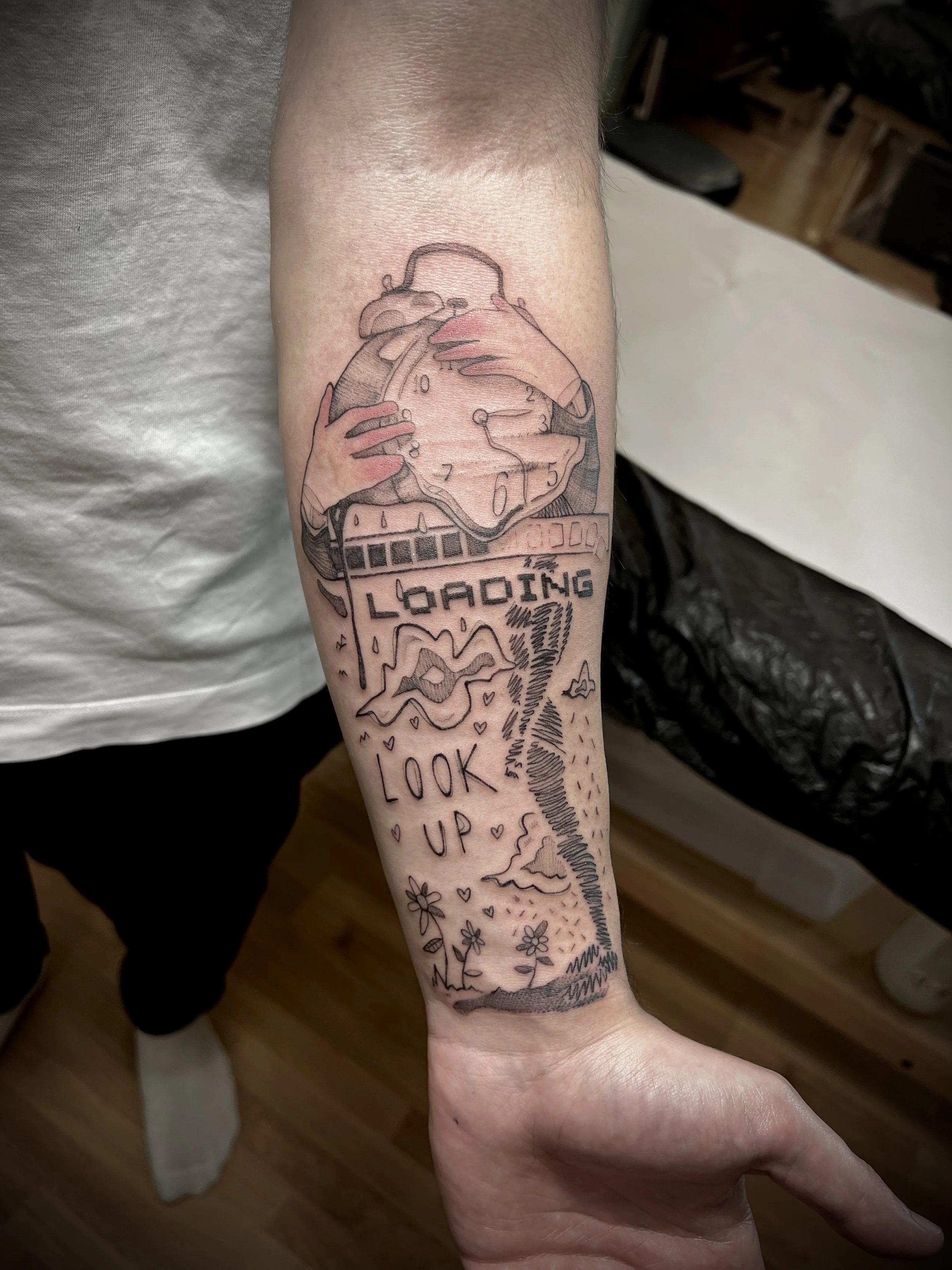 Tatuaggio sul braccio con un orologio d'epoca, una manina che lo tiene, una schermata di caricamento con la scritta 'Loading', e le parole 'Look Up' circondato da fiori, cuore e nuvole.