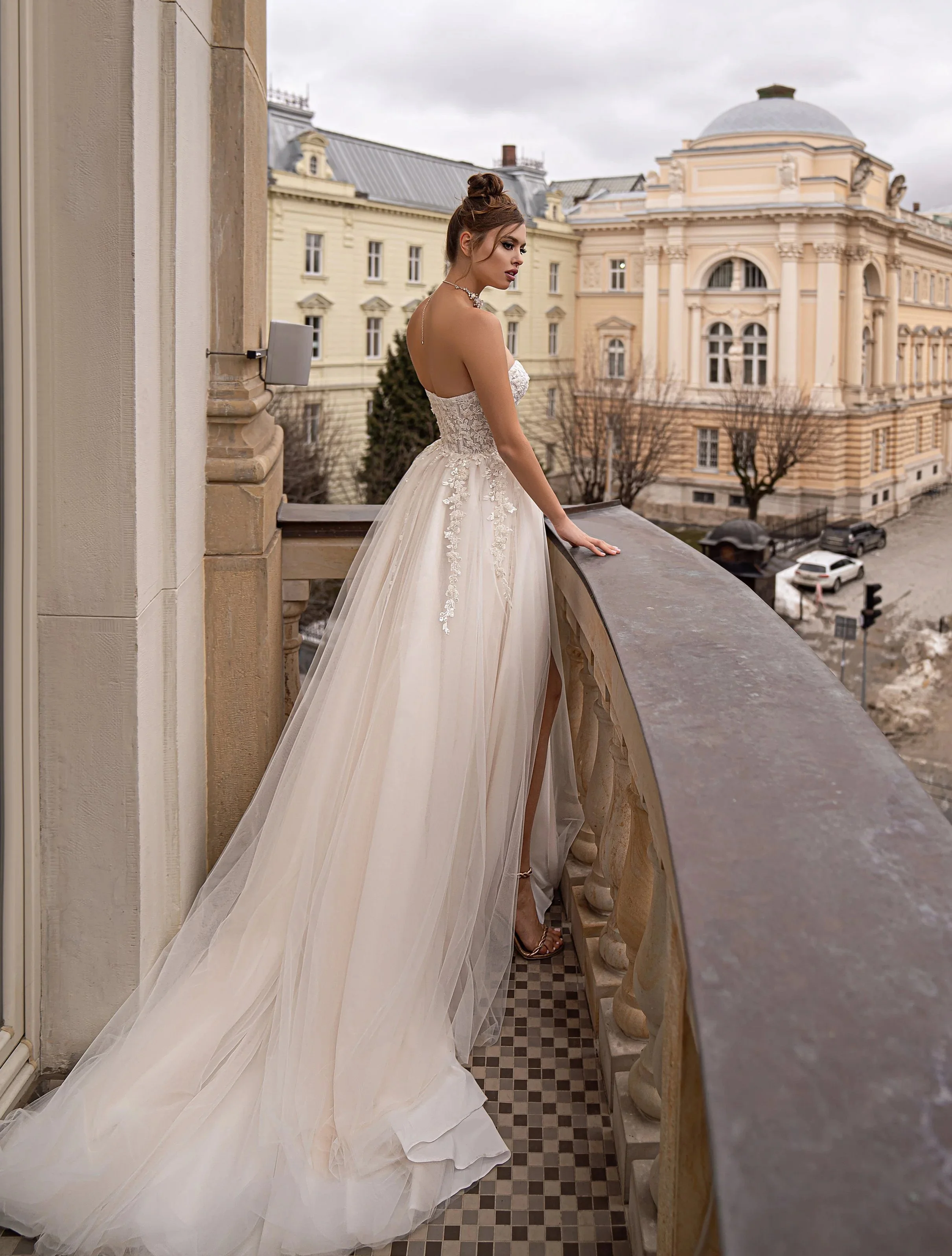 Femme en robe de mariée élégante sur un balcon en ville, avec des bâtiments historiques en arrière-plan, un ciel nuageux.